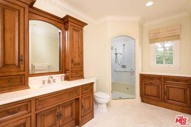 这间主浴室的米色墙壁、天花板和地板都装饰着水槽区和梳妆台区的红木橱柜和抽屉，它们都有白色的台面，与白色的马桶相匹配。