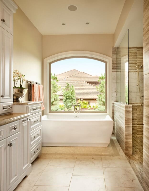 淋浴区旁边的独立式浴缸由一扇白色框架的大拱形玻璃窗照亮，玻璃窗的背景是米黄色的墙壁和米黄色的大理石地板。