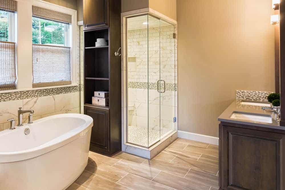 这间可爱的浴室有一个靠窗的独立式浴缸，浴室头部有一个小木柜子，靠近带有两个洗手池的梳妆台对面的步入式淋浴间。
