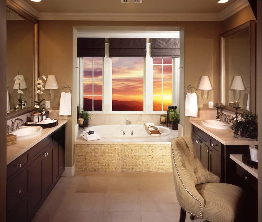 白色窗户外的日落与主浴室的米色元素形成了鲜明的对比，主浴室两侧有两个梳妆台，两侧是浴缸的凹室，侧面有米色瓷砖。