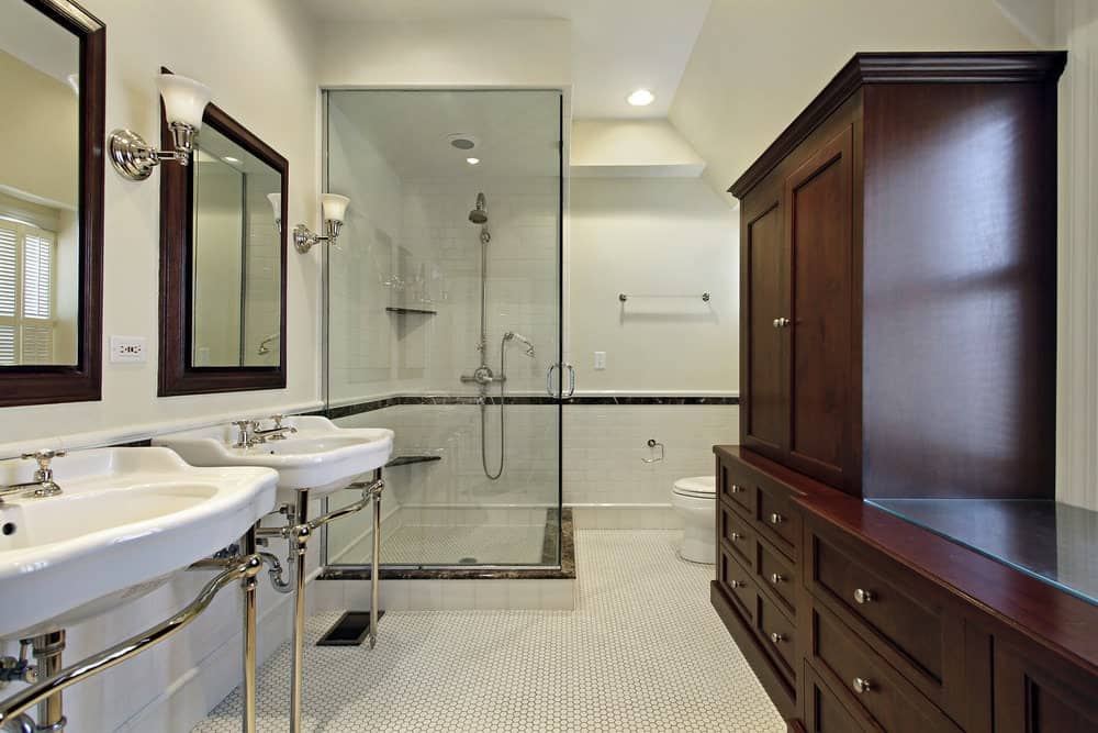 白色的瓷水槽由银架支撑，顶部有木框镜子，与白色厕所旁边的木结构橱柜和抽屉很相配。