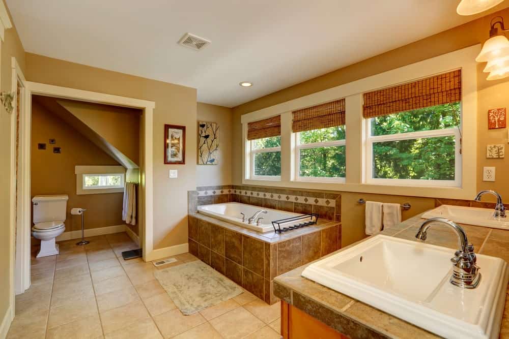 墙壁和地板的泥土色调与浴缸镶嵌的棕色大理石瓷砖相辅相成，洗手池区域的台面由壁挂灯的黄色灯光照亮。