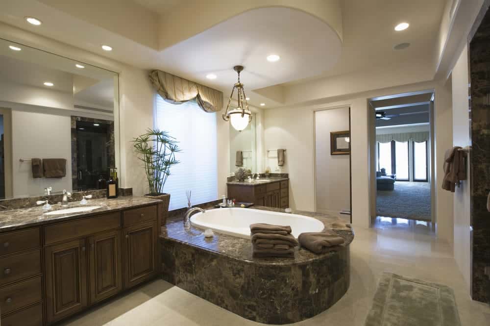 米黄色地板中间的深色大理石浴缸与梳妆台的台面相匹配，梳妆台有优雅的固定装置，一面嵌入米黄色墙壁的大镜子。