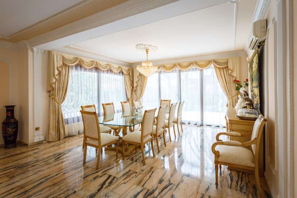宽敞的餐厅，瓷砖地板和全高玻璃穿着优雅的窗帘和帷幔。它包括一个水晶吊灯和玻璃餐桌，配以黄色软垫椅子。