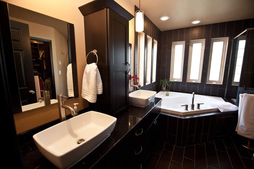 黑色浴室拥有一个浴缸和一个带镜子的双容器水槽梳妆台，中间还有一个深色的木质储物柜。它由玻璃吊坠和平顶天花板灯照明。