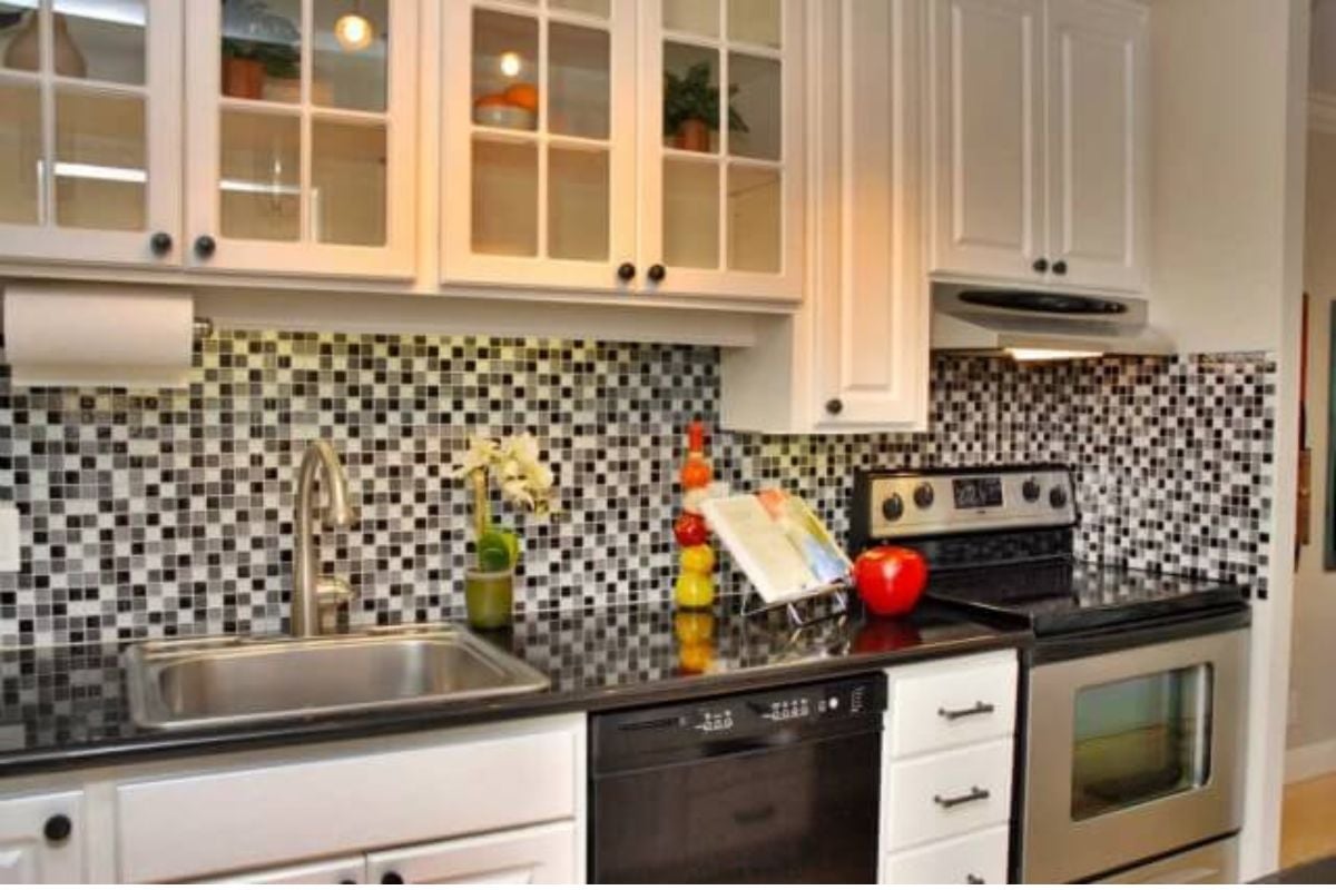 一间白色厨房，主准备区和水槽上方有几个玻璃柜。后挡板是混合黑色，灰色和白色的马赛克玻璃瓷砖。