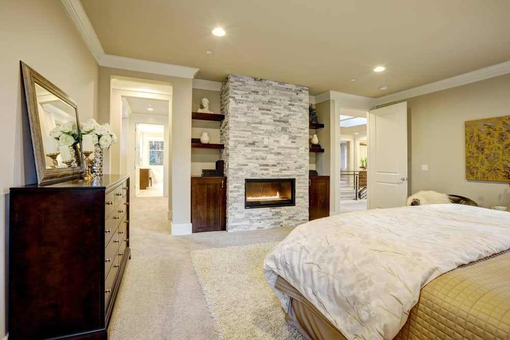 米色的主卧室设有白色的皇冠造型和地毯地板，上面有蓬松的地毯。卧室里有一座壁炉，壁炉安装在正对着床的一面石砖墙上。