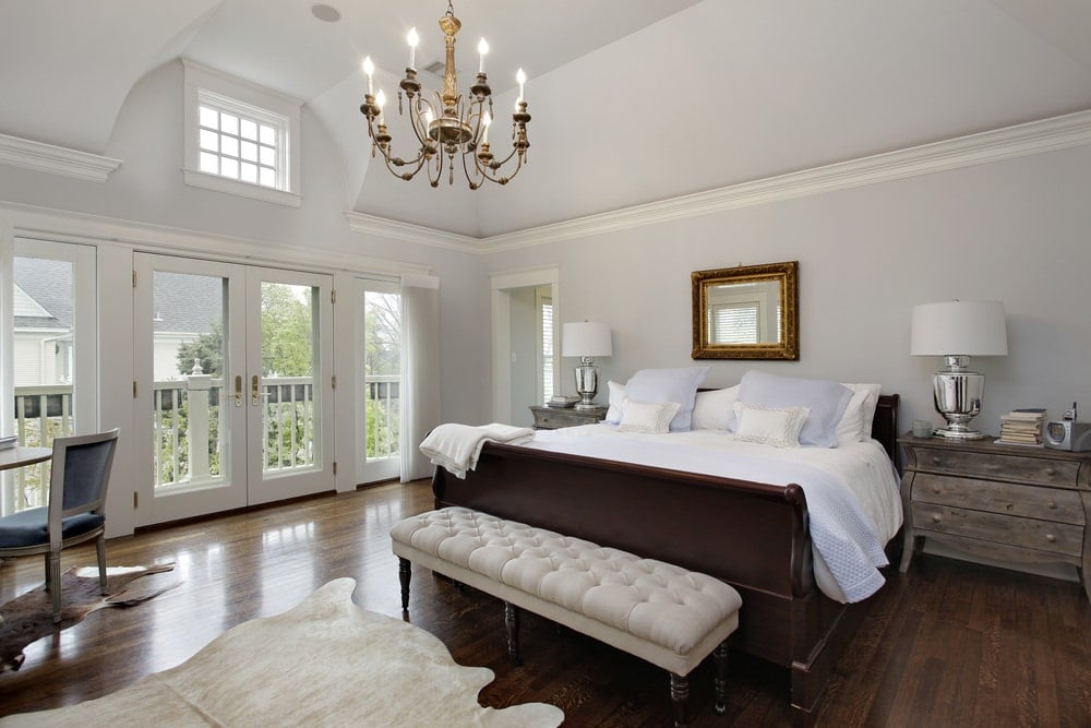 白色的主卧室，有一盏蜡烛吊灯，硬木地板上铺着牛皮地毯。它包括乡村风格的床头柜和一张深色木床，床的一端有一张米色的簇绒长椅。
