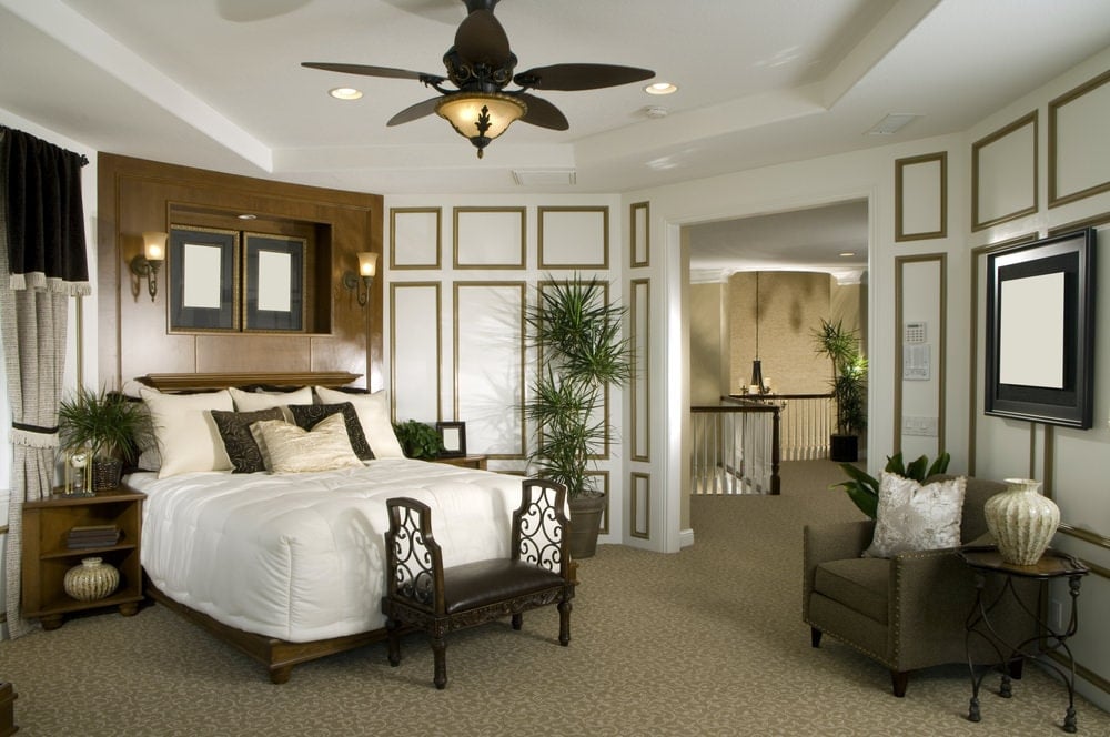 热带的主卧室，有全高的护壁板和托盘天花板，安装有嵌入式灯和一个带灯的青铜吊扇。它包括一张木床、一把灰色扶手椅和一个圆形金属边桌。