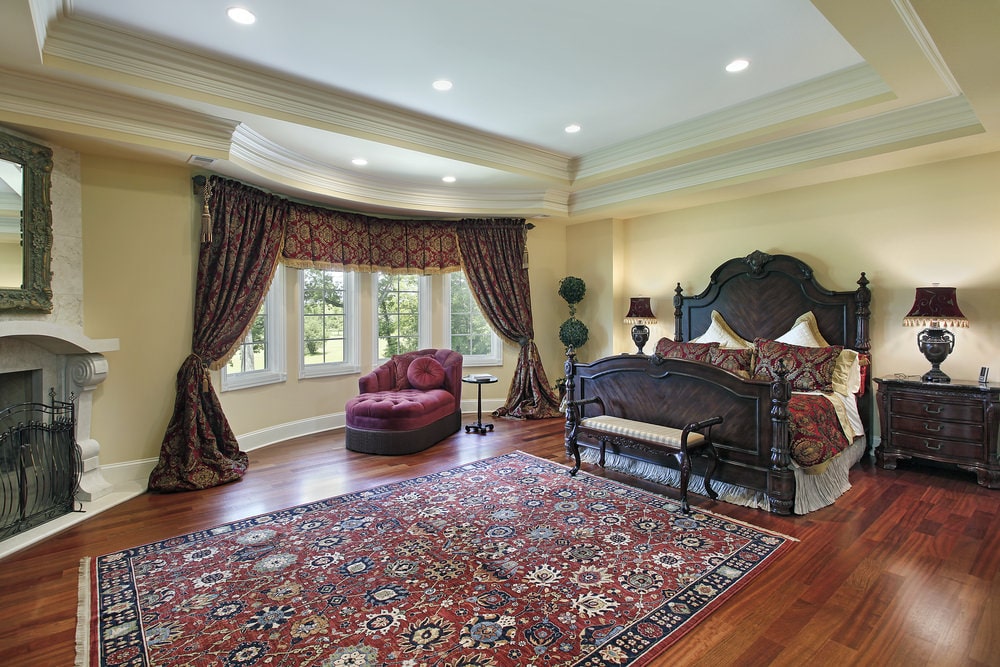 精致的主卧室展示了一张深色的木床和一个红色的簇绒躺椅，在凸窗边装饰着经典的窗帘和帷幔。它包括一张印花地毯和一个壁炉，上面有一面华丽的镜子。