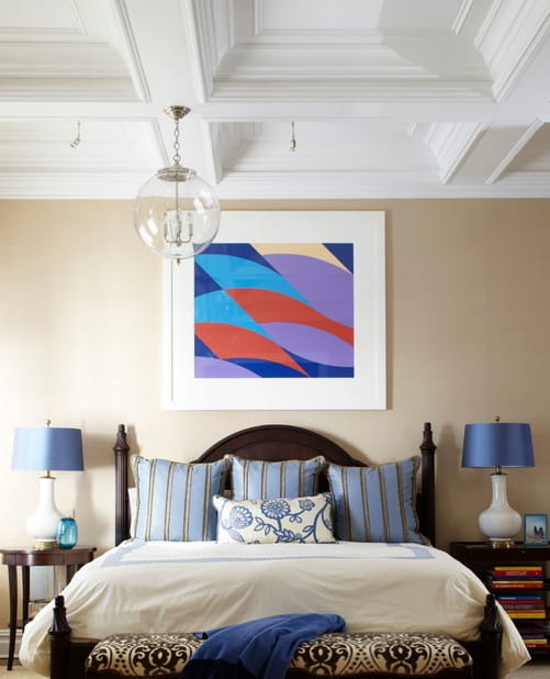 这间主卧有一张舒适的床和木制床头柜，上面有蓝色的灯罩，一件抽象艺术品为这间主卧带来了一抹色彩。它被挂在格子天花板上的玻璃球形吊坠照亮。