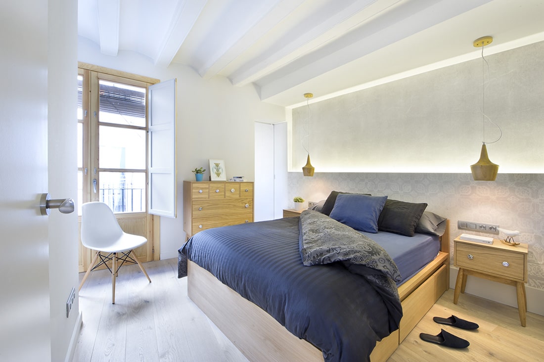 白色的主卧室，宽阔的木板地板和桶形拱形天花板，上面安装着木制吊灯。它包括一把现代的白色椅子和一张舒适的床，两侧是与梳妆台相匹配的浅色木制床头柜。
