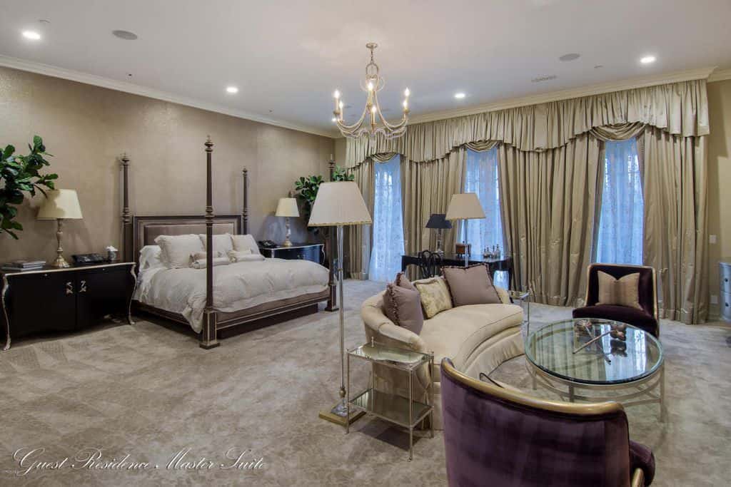 豪华的主卧室，天鹅绒地毯地板和全高窗户穿着优雅的窗帘和窗帘。它包括一张四柱床和一个由蜡烛吊灯和嵌入式顶灯照明的休息区。