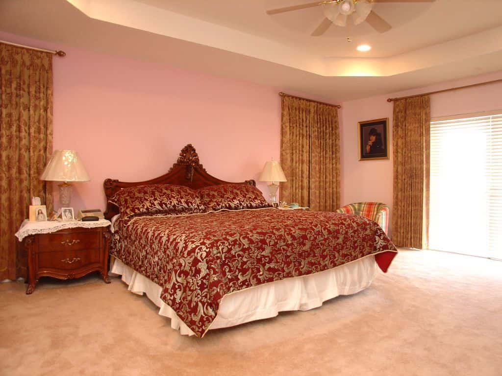 优雅的主卧室，地毯地板和托盘天花板安装了嵌入式灯和白色风扇。它包括一张条纹圆靠背椅和一张裹着优雅图案床上用品的裙床。