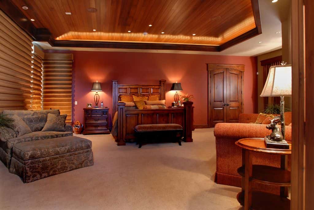 温馨的主卧室配有舒适的座椅和木床，床头柜上配有时尚的台灯。它有地毯地板和覆盖着深色木板的托盘天花板。