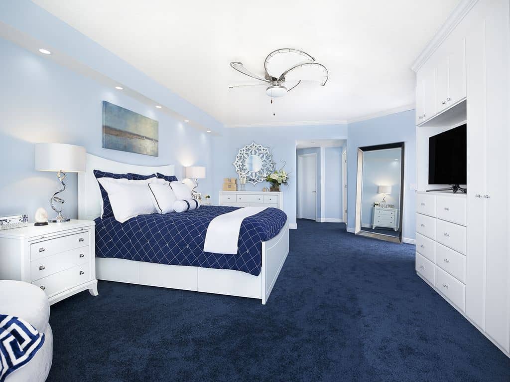 白色梳妆台上方挂着一面华丽的日晒镜，正对着蓝色地毯地板上光滑的床。它的两侧有三个有抽屉的床头柜和时尚的台灯。