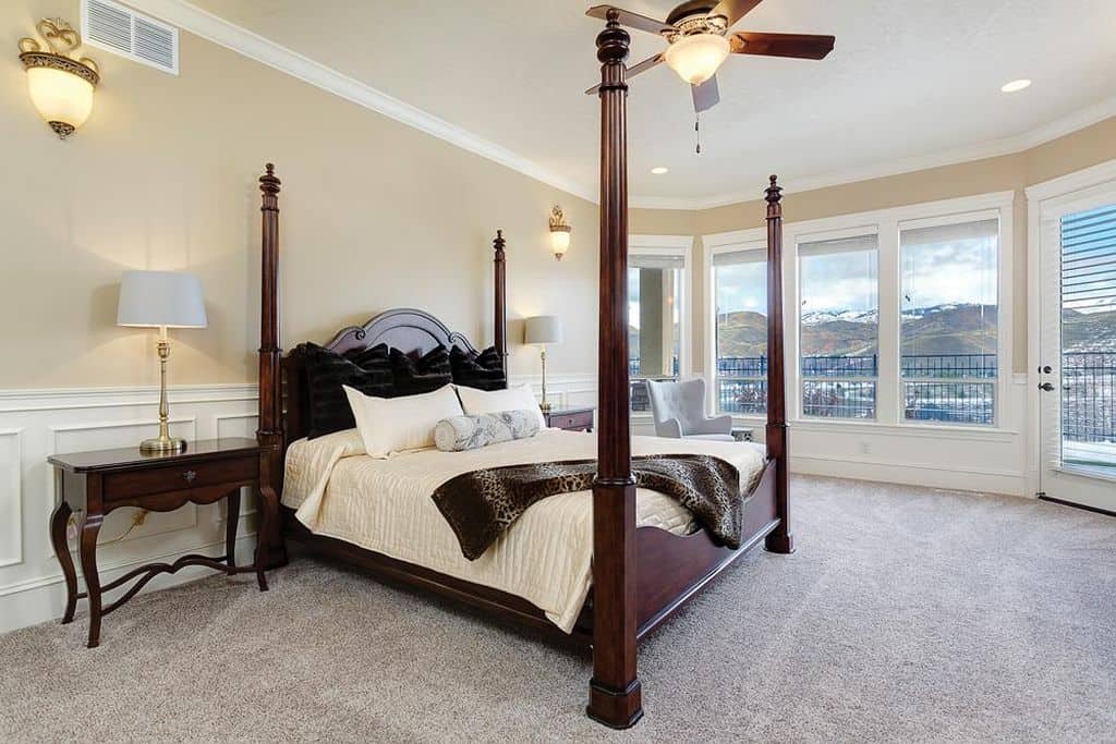 精致的主卧室，配有灰色地毯地板和玻璃板窗户，俯瞰着壮丽的山景。它包括一把羽绒靠背椅和一张四柱床，两侧配有配套的床头柜和黄铜台灯。