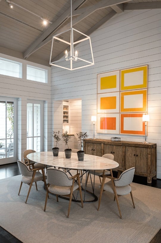 充满活力的橙色墙壁艺术为这个餐厅增添了温暖的口音，展示了一个白色的立方体吊坠和优雅的餐厅设置在质朴的自助餐桌上，上面有灯罩。