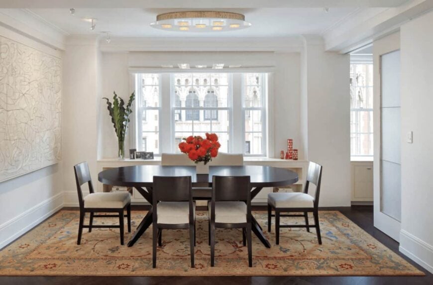深色木质椭圆形餐桌在白色餐厅中形成了鲜明的对比，在经典的区域地毯上提供软垫椅子，玻璃窗旁设有内置橱柜。