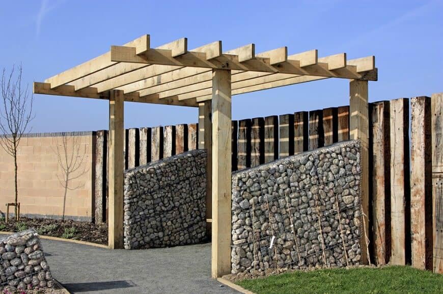 一个与乡村木栅栏的外观相匹配的凉棚。柱子之间的堆叠石头增加了风格和流畅的运动，从走道到栅栏。