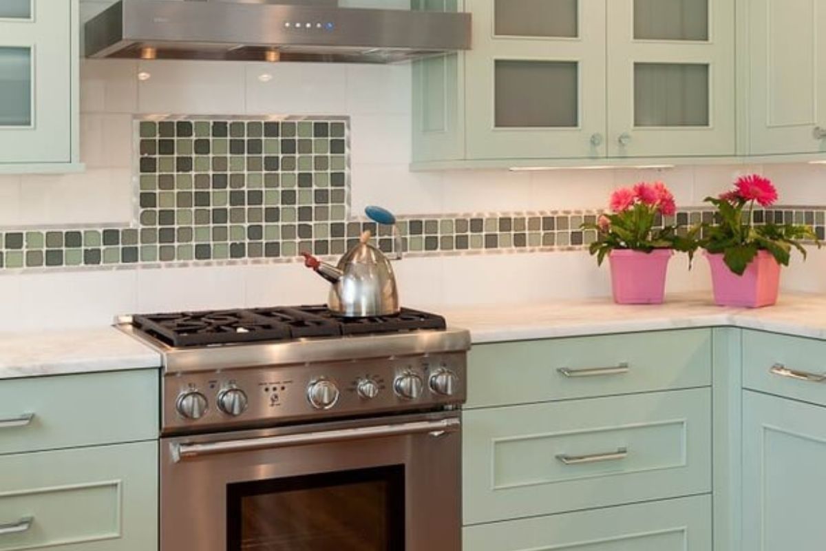 这是一间简单而温馨的乡村厨房，配有薄荷味的橱柜，白色瓷砖后挡板上的绿色瓷砖图案交替出现。
