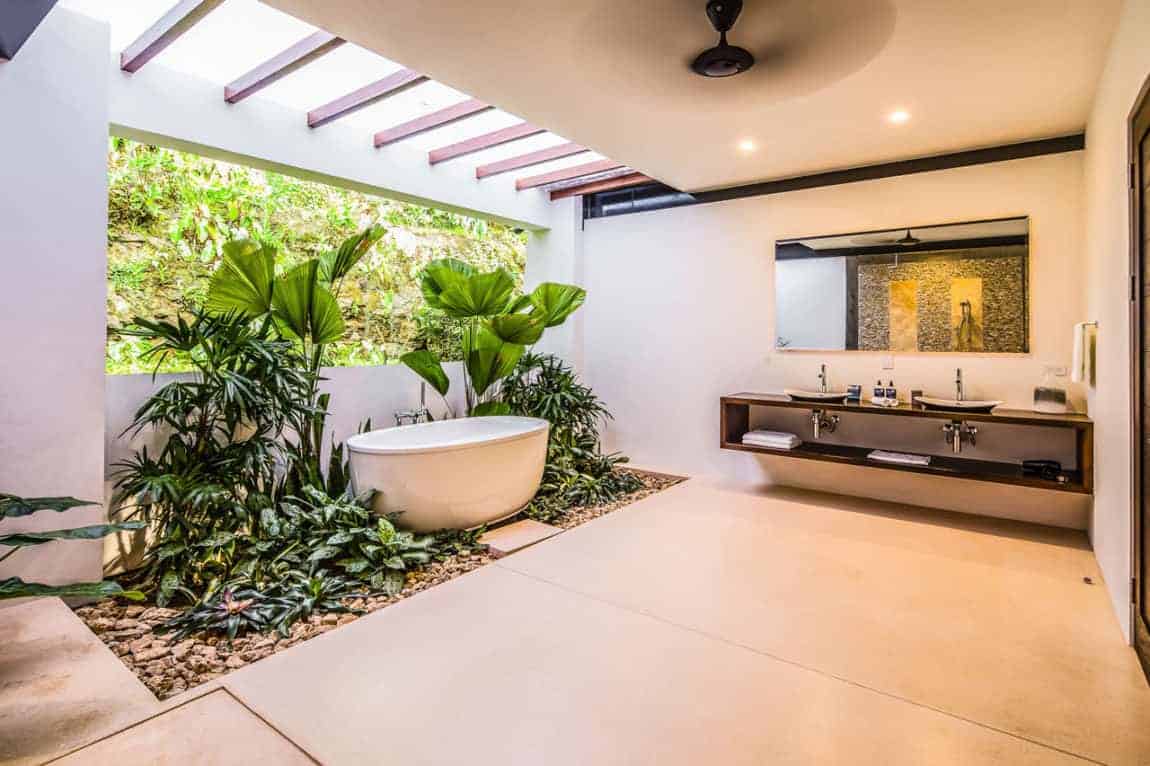 这个华丽的热带风格浴室有一个微型热带花园，围绕着独立的浴缸，其区域被上方天窗和后面宽大的窗户倾泻而下的自然光照亮。它还配有一个木制浮动梳妆台和两个简单的水槽。