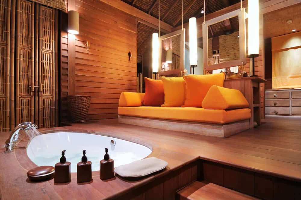 这是一间宽敞的热带风格浴室，木质教堂天花板与木质墙壁相匹配，与远处墙上的木质梳妆台融为一体。这是由一个带有橙色靠垫和枕头的木制长凳与下沉的浴缸区分开。