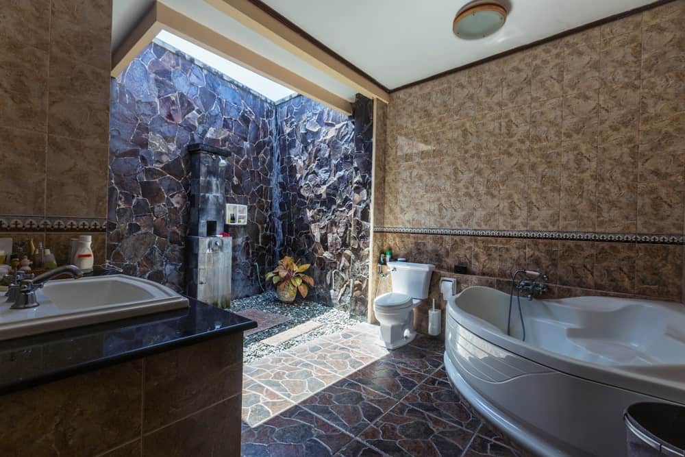 卫生间旁边有一个大浴缸，在棕色大理石墙壁和花纹地砖的映衬下显得格外显眼。这个浴室的亮点是室外淋浴区被高质感的石墙包围，自然光线增强了淋浴区的效果。