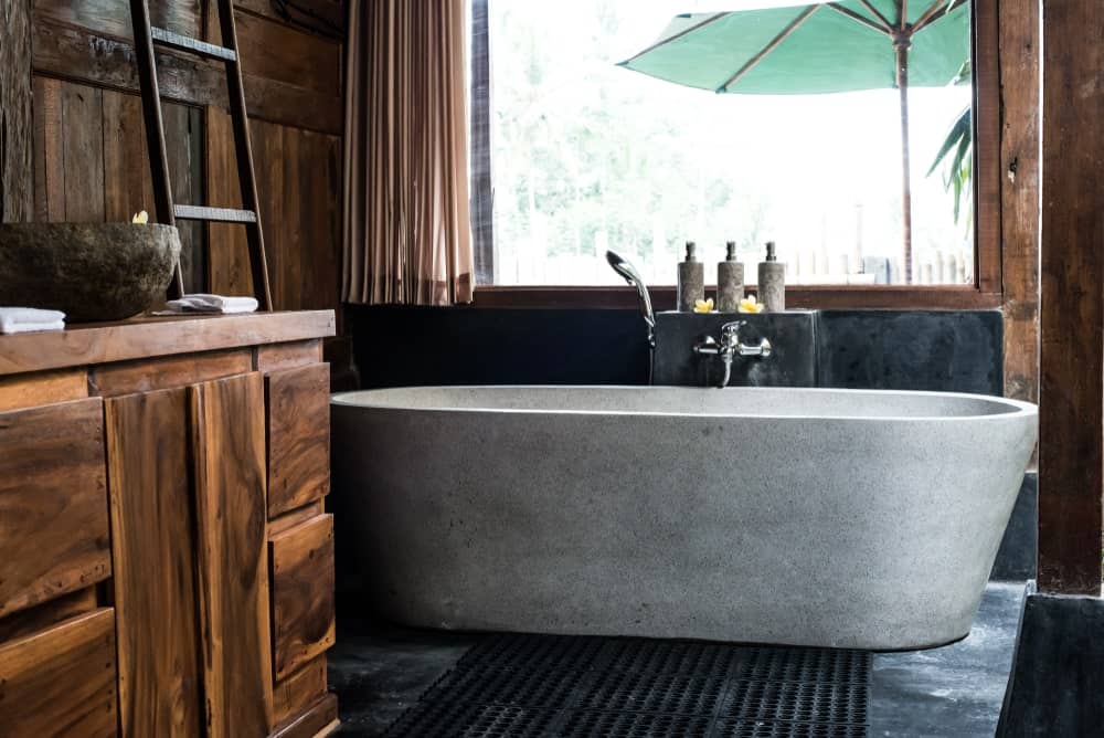 这间简单的热带风格浴室有一个灰色混凝土独立式浴缸，与深色地砖和后挡板很好地搭配，木墙与浴缸旁边的木制梳妆台融合在一起，浴缸旁边有一个有质感的棕色洗手池。