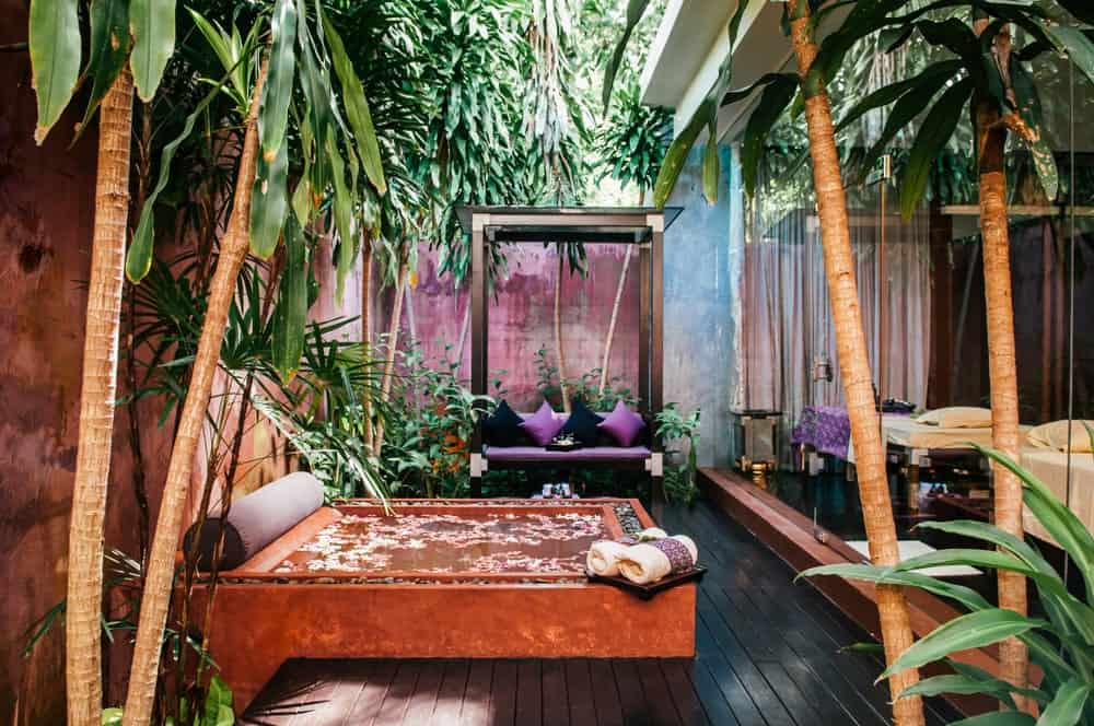 这个热带风格的浴室有一个被热带树木和植物包围的浴缸，让你感觉像在森林里的池塘里洗澡，让你免受阳光的照射。旁边还有一个放松的休息区，有紫色的靠垫和枕头。