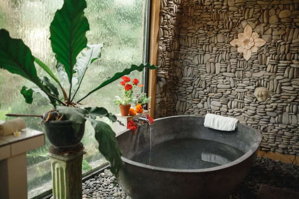这个热带风格浴室的圆形石头浴缸与鹅卵石地板以及后面由各种石头制成并装饰有花卉艺术品的墙壁很好地搭配。这些都被高高的玻璃窗照亮了，为了保护隐私，流水模糊了窗户。