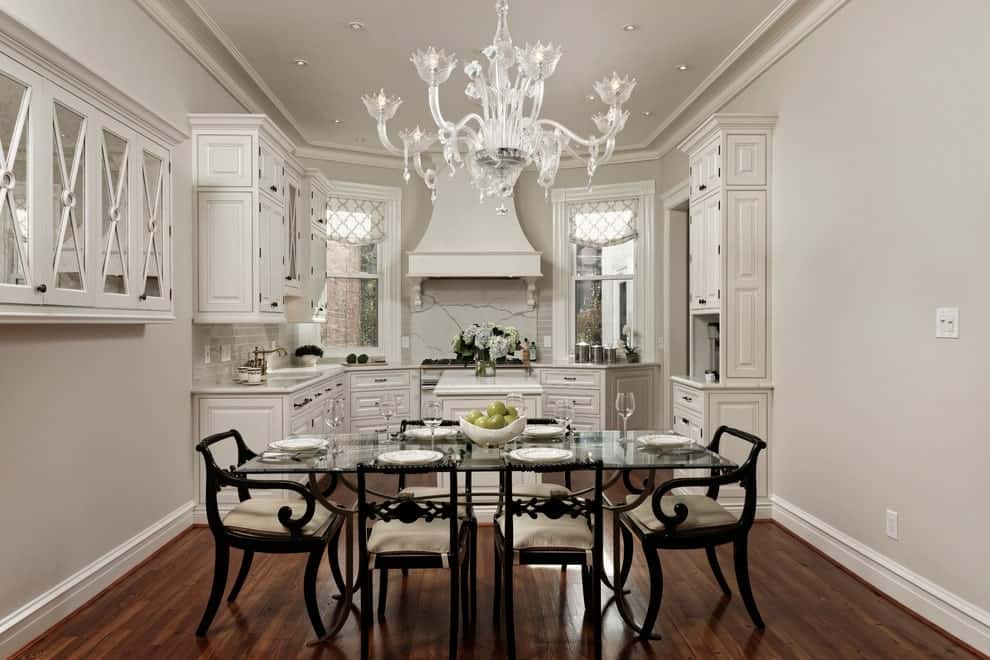 白色厨房与共享用餐空间，设有深色木质软垫椅和由华丽的玻璃吊灯照明的矩形餐桌。屋内铺着厚实的硬木地板，还有装饰着罗马窗帘图案的落地窗。