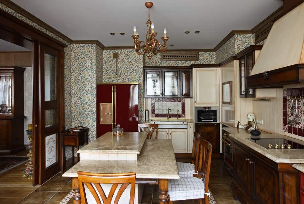 这间厨房贴着碎花壁纸，米黄色的后挡板配以红边瓷砖。它有木制橱柜和一个凸起的早餐岛，上面有格纹软垫椅，由一盏经典的枝形吊灯照亮。