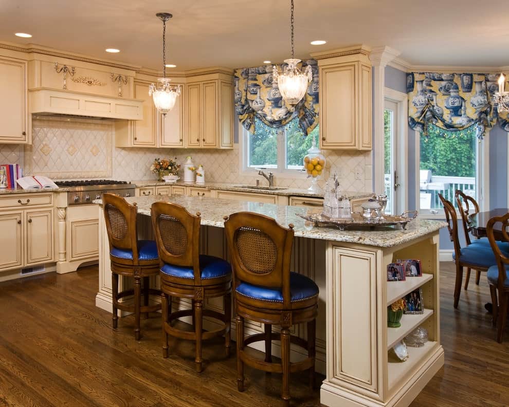 维多利亚式厨房，丰富的硬木地板和玻璃窗户覆盖花瓶印刷罗马窗帘。它包括米色的橱柜和花岗岩顶部岛台，配有配有蓝色靠垫的圆形柜台椅。