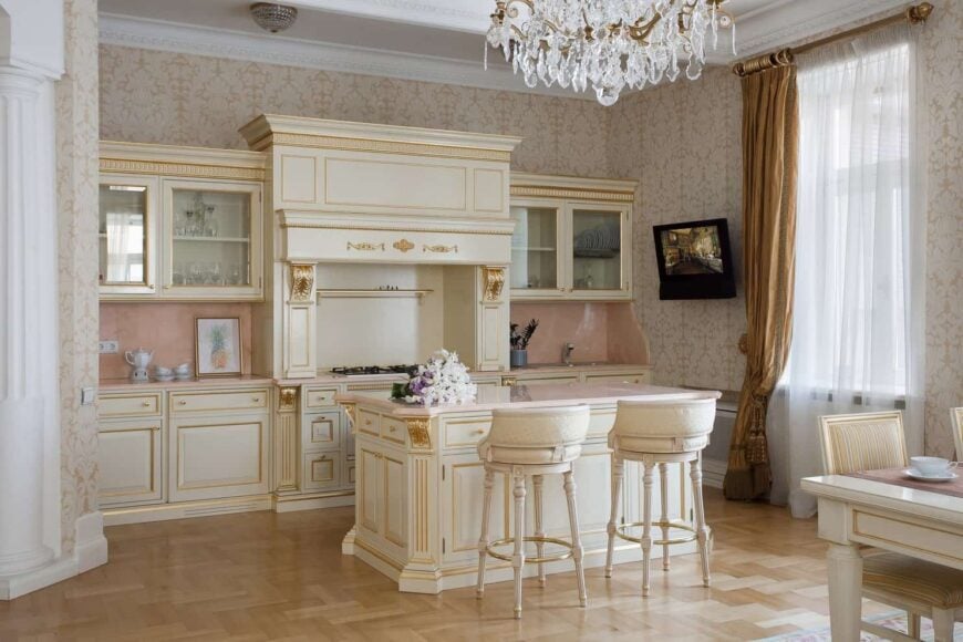 这个厨房拥有精致的花纹壁纸，奶油色橱柜和配套的早餐岛，配有金色装饰。它有浅色的硬木地板和玻璃窗，上面覆盖着白色透明窗帘和优雅的棕色窗帘。