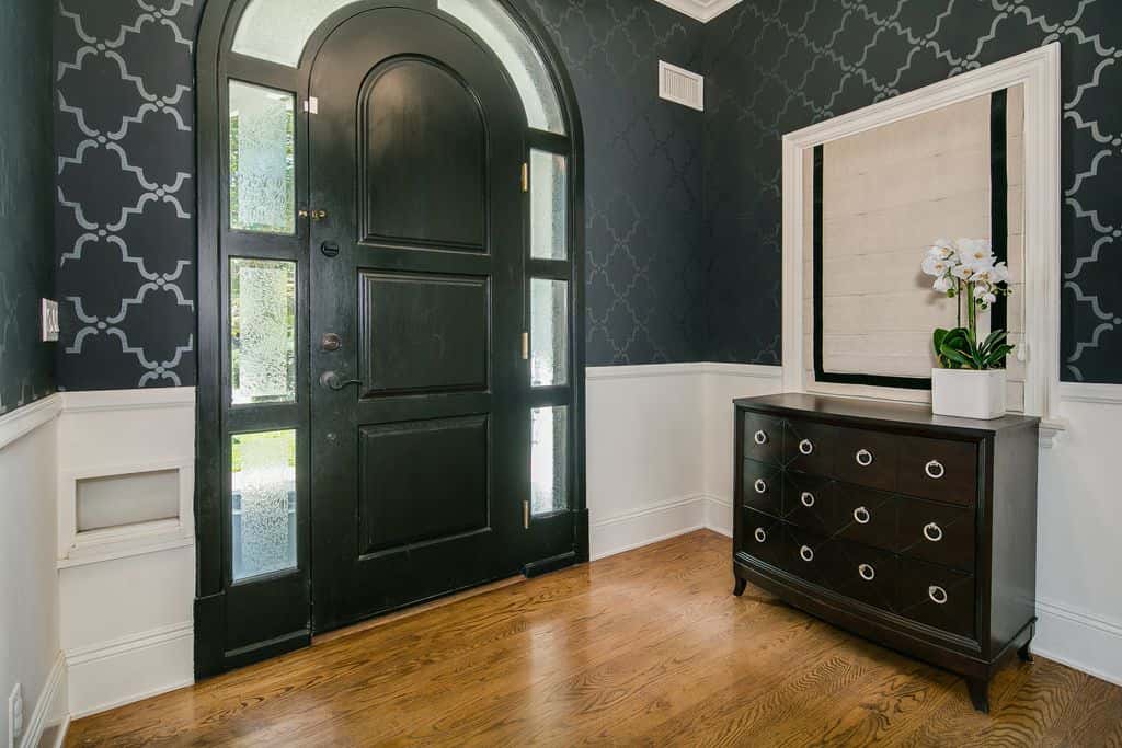 这个门厅展示了白色护壁板上方的半图案黑色墙壁。它有一扇黑色的拱形门，周围环绕着玻璃板，毗邻一面白色镜子下的木制控制台桌子。