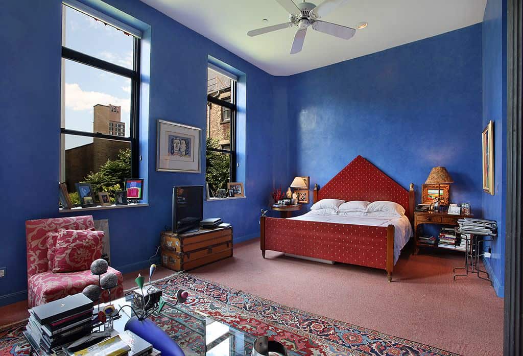 这间蓝色的主卧室与一张红色点床和一把有图案的椅子形成了美丽的对比，椅子正对着经典地毯上的玻璃咖啡桌。它有地毯地板和充满活力的蓝色墙壁，装饰着可爱的艺术品。
