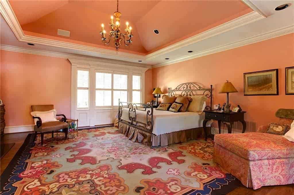 一盏老式的枝形吊灯悬挂在华丽的金属床上，照亮了这个珊瑚粉色的卧室，配有碎花躺椅和棕色扶手椅，扶手椅放在可爱的地毯上。