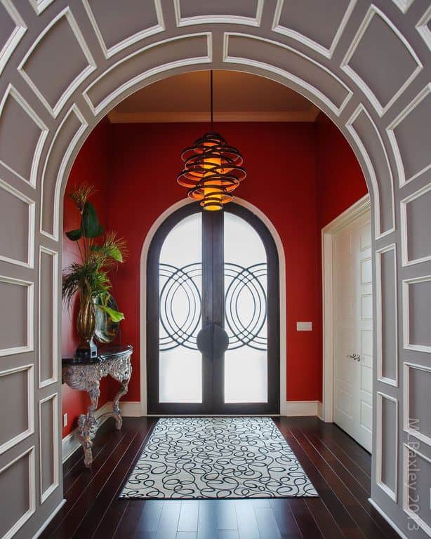 灰色的拱门覆盖着护壁板，通往红色的门厅，带有法式门和装饰华丽的控制台桌子，由现代吊灯照明。它有深色的木板地板，上面有白色的漩涡地毯。