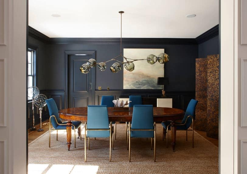 围绕木桌的蓝色餐椅靠垫在黑色墙壁的映衬下显得格外突出，墙壁上有一致的黑色护墙板。
