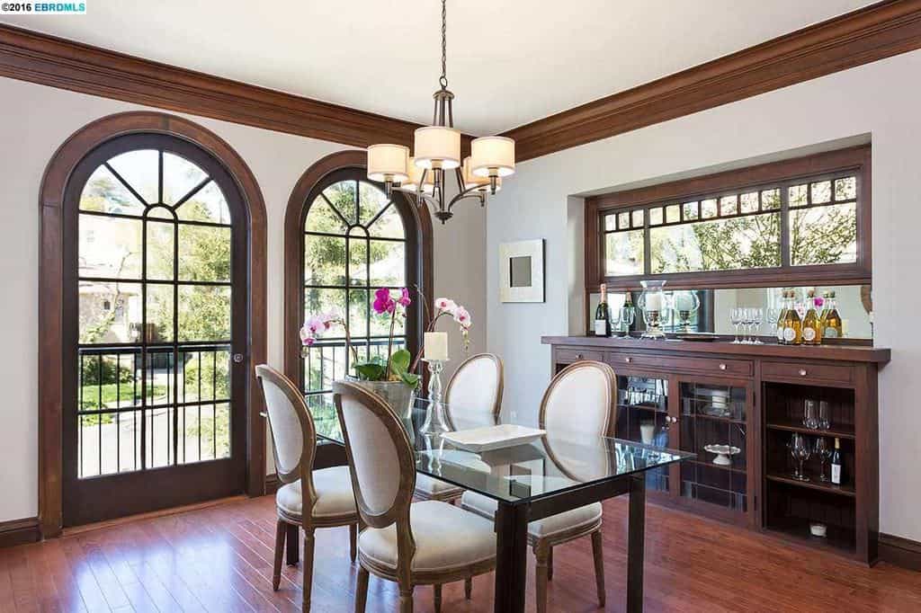 带有拱形顶部的玻璃法式双开门为玻璃桌面和白色软垫椅子提供照明。门的框架与天花板的线条和一扇矩形窗户下的内置橱柜相匹配。