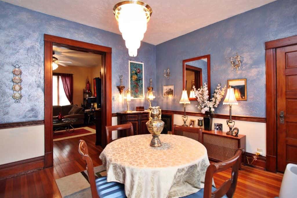 明亮的蓝色墙壁有一种闪闪发光的效果，与橱柜、门、门框和椅子的深色木质元素完美地互补，圆桌周围的桌布在天花板上安装的半平光灯的黄色灯光下闪闪发光。