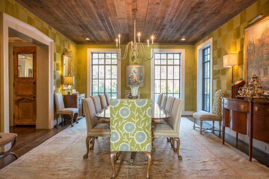 黄绿色的墙壁上有一种微妙的格子图案，与长木制餐桌周围的软垫椅子上的花卉图案很好地搭配在一起，餐桌上有一盏细黄铜枝形吊灯。