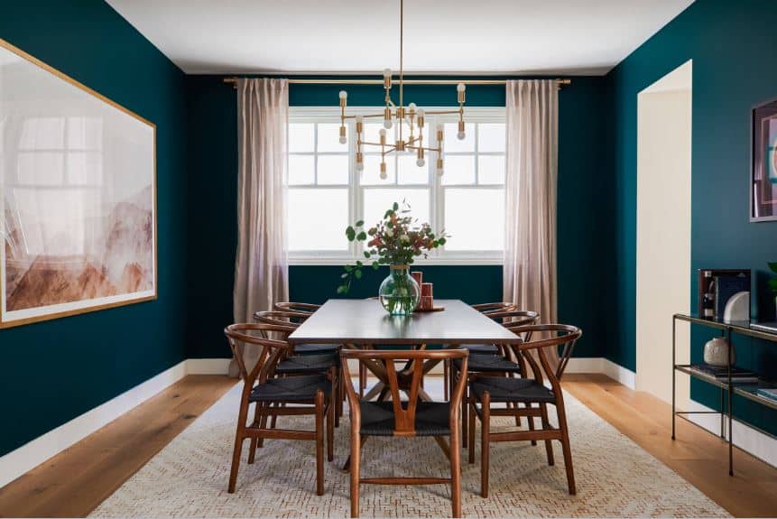 木制餐桌周围环绕着木制叉骨椅，椅子上有绿色编织的柳条座椅，与深绿色的墙壁相呼应，桌子旁边安装着一幅大型艺术品。