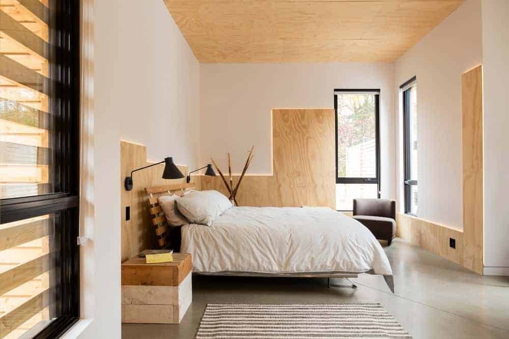 这间主卧室的天花板由裸露的胶合板制成，与白色墙壁的木质基调相匹配，并配有背光照明，以强调线条。工业风格的混凝土地板与木床相得益彰，木床两侧是重新利用的木制调色板盒。
