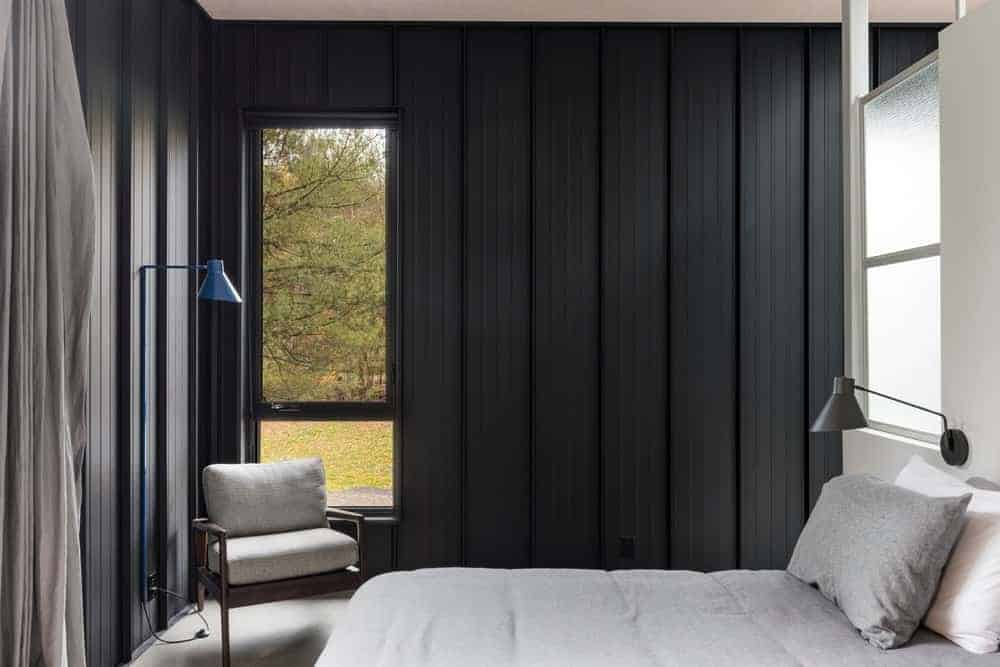 浅灰色的床单与灰色的混凝土地板和窗边的木制扶手椅的靠垫相匹配，扶手椅镶嵌在深灰色材料的工业风格墙壁上。