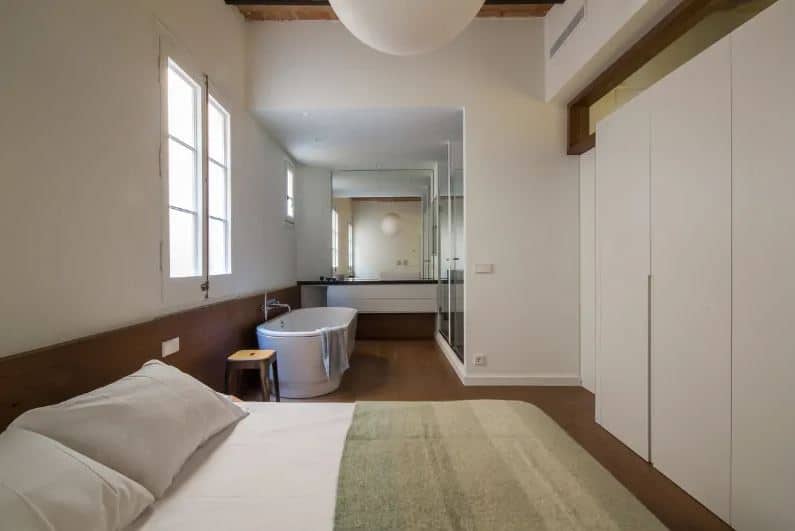 这是一间工业风格的卧室，挨着浴室，铺着同样的硬木地板，白色的靠垫与没有床架形成对比。硬木地板延伸到较低的护墙板，与露出木梁的木质天花板相匹配。