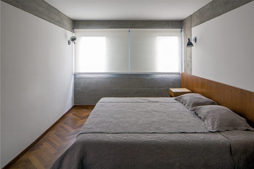 白色的墙壁和天花板采用了灰色混凝土，与灰色床单相匹配，床上的木制床头板延伸到床头柜，与硬木地板相匹配。