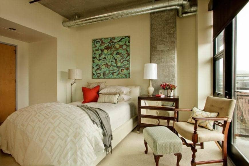 这个迷人的主卧室是传统风格和工业风格的可爱融合。它有一张传统的床，与米色的地毯地板和米色的墙壁相得益彰。暴露的管道和混凝土天花板的通风口突出了这一点。
