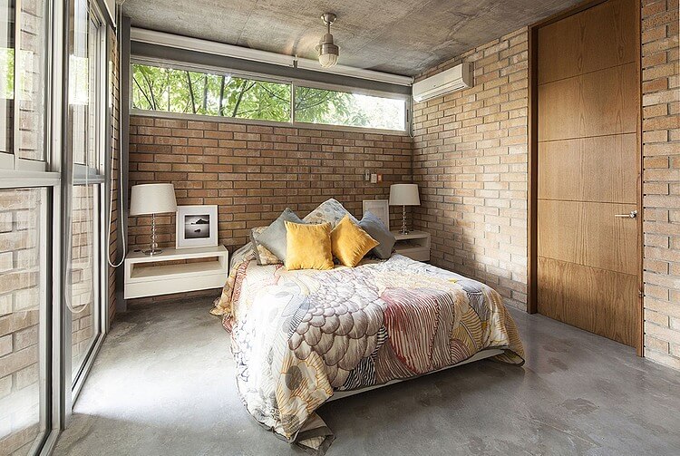 这间工业风格卧室的灰色混凝土地板与灰色混凝土天花板相匹配，天花板支撑着床上半平装的灯，彩色床单与红砖墙相得益彰。